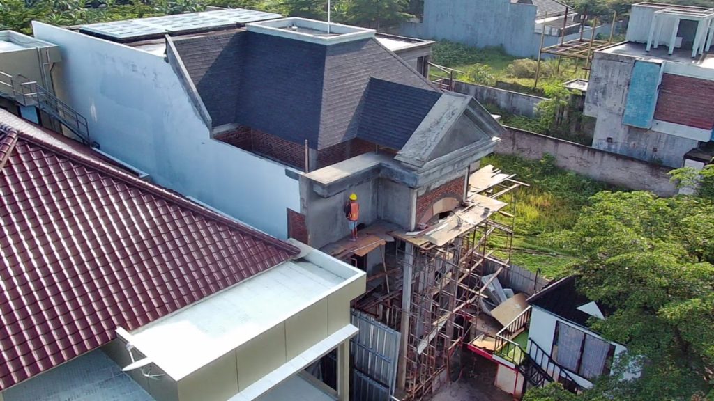 Tampilan aerial proses pembangunan rumah bergaya klasik, memperlihatkan detail atap limasan dan elemen fasad depan yang sedang dikerjakan oleh pekerja konstruksi.